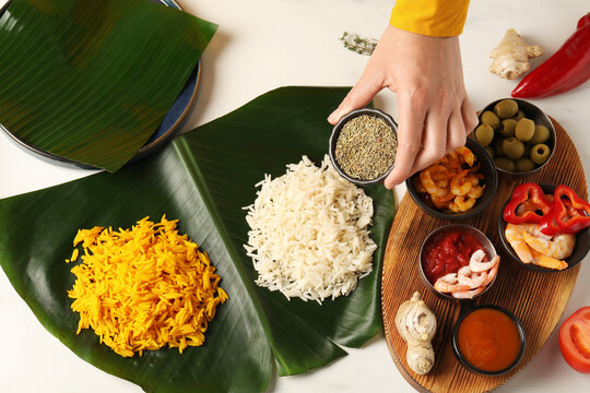 Woman holding bowl of cumin at white table with banana leaves and tasty food, top view