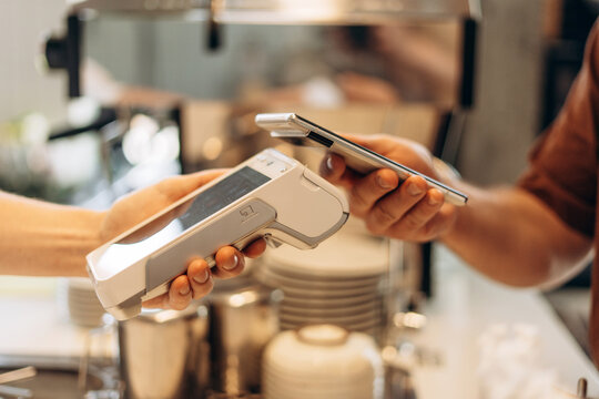 Customer is holding his smartphone over a payment terminal, making a contactless payment for his coffee at the counter of a coffee shop