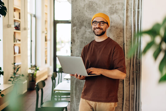 Smiling happy middle aged Latin freelancer man wearing yellow hat stylish eyeglasses, holding laptop