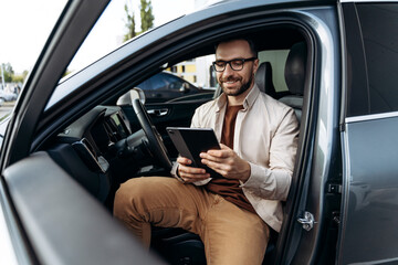 Happy, smiling Latin man holding digital tablet, using mobile app, sitting in modern new car