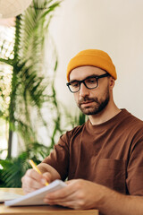 Pensive bearded man wearing stylish eyeglasses and yellow hat taking notes, working at workplace