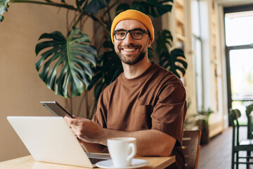 Smiling, bearded man wearing stylish yellow hat, businessman holding digital tablet, using laptop