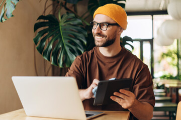 Portrait of smiling, happy man, hipster wearing yellow hat holding digital tablet wipe, using laptop