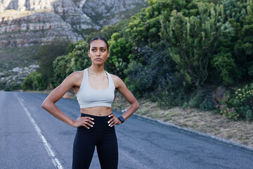 Confident female runner standing on an abandoned road and looking away. Slim woman in fitness attire with hands on hips relaxing during a run in the park.