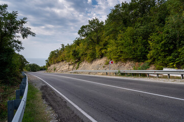 Road landscape forest, mountains and sea on the horizon. Empty long mountain road to the horizon on a sunny summer day with dramatic cloudy sky. Highway turn panoramic view.