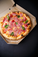 Pizza with cheese, ham, green basil leaves and tomatoes in paper box on black background. Food product advertising banner, copy space