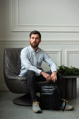 Fototapeta premium A man is comfortably sitting in a chair, resting with a leather backpack near him