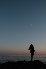 Fototapeta premium Woman alone standing on the coast overlooking the sea and sky. Woman silhouette in the dusk sky under the crescent moon.