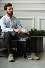 A man is comfortably sitting in a chair, resting with a leather backpack near him