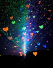 夜空に放たれる虹色の光の束が、多彩な色の輝くハートに囲まれ、空中に漂うように見えます。暗い風景と鮮やかな色彩の対比が、幻想的でロマンチックな雰囲気を生み出しています。






