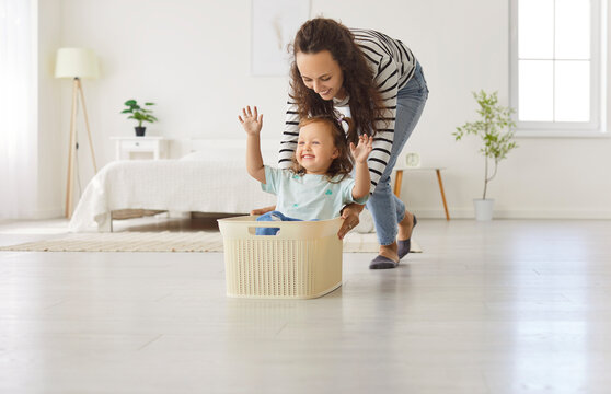 Young happy smiling mother having fun in the living room at home together with little cute child girl riding her in toy basket. Smiling female parent spending time with daughter playing together.