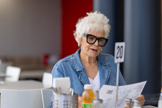 elderly lady wearing denim and thick rimmed black glasses sitting at cafe table looking at menu