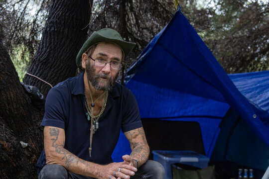 homeless man sitting under trees where he has set up a tent for shelter