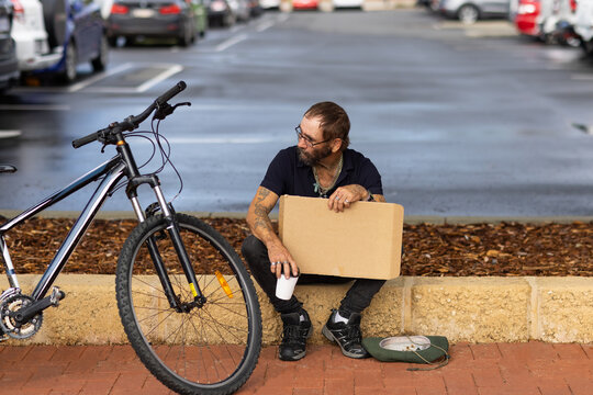 middle-aged homeless man sitting on verge with blank sign and hat down on footpath  asking for help