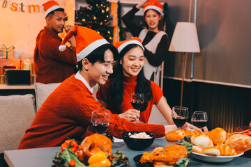 Group of young Asian man and women as friends having fun at a New Year's celebration, holding gift boxes standing by Christmas tree decoration, midnight countdown Party at home with holiday season.