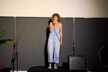 Caucasian female performer with curly hair delivering a monologue on stage in a dimly lit theater...
