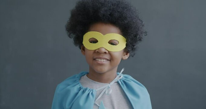 Slow motion portrait of creative African American kid in superman mask and cape smiling looking at camera on gray background. Childhood and game concept.