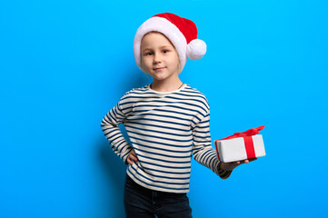 Happy new year concept. Caucasian preschool girl wearing santa claus red hat holds christmas gift on blue isolated background