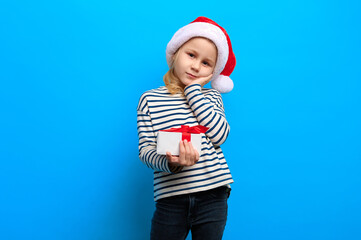 Happy new year concept. Caucasian preschool girl wearing santa claus red hat holds christmas gift on blue isolated background