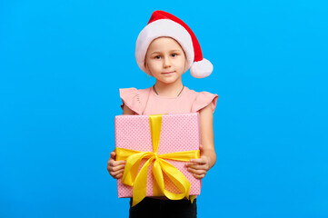 Happy new year concept. Little white girl in santa claus red hat holds christmas pink gift box with yellow bow on blue isolated background in studio