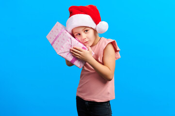 Excited girl in a Santa hat with oversized glasses presents a pink ribbon-wrapped gift against a bold blue backdrop. Radiates festive charm and holiday cheer...