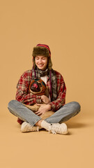 Smiling young man in hat and checkered jacket sitting with happy pet, Pug dog in knitted hat, with tongue sticking out, against studio background. Concept of animal, winter season, friendship, fashion