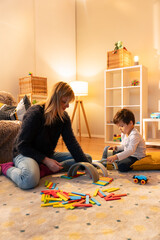 Childminder and toddler playing with colorful wooden building blocks on a rug in playroom