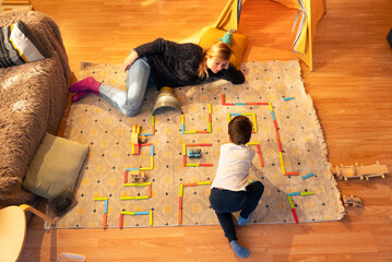 Child playing with colorful wooden toy road on carpet with mother relaxing nearby