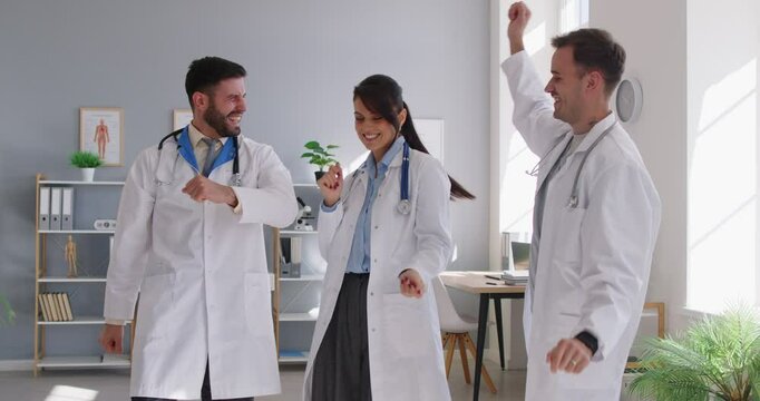 Team of happy doctors celebrating success having fun together. Group of 3 medical professionals give each other high five and dance in staff room celebrating promotion, pay raise or patient's recovery