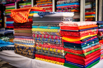 Stacks of colorful fabrics displayed at an Indian market, featuring intricate patterns and vibrant textiles, showcasing traditional craftsmanship.