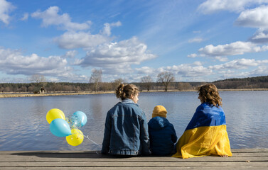 three people, adult and child, near lake on sunny spring day, holding yellow and blue balloons in...