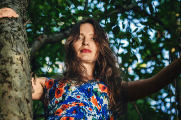 Fototapeta premium Bottom view of mature smiling woman with long brown hair on tree. Looking at camera. Blurred background.