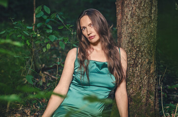 Fototapeta premium Relaxed woman, resting near tree with rough trunk and bark, looks into camera, tilting head to side.