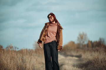 Teenage girl with red hair and glasses standing in a windy field, with hair blowing, dressed warmly in a brown coat and pink sweater.