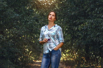 Woman in shirt and jeans looking up, holding glasses, standing on trail in dark green forest. Walk.