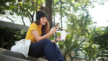Gen Z eco go green woman asia sit happy relax at tree public park city life enjoy typing mobile...