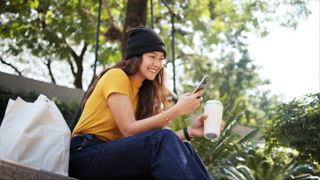 Gen Z eco go green woman asia sit happy relax at tree public park city life enjoy typing mobile phone drink coffee bottle use reuse tote bag care save the earth planet net zero waste clean future hope