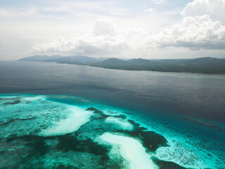 An aerial view of a vibrant coastal lagoon with coral reefs and turquoise waters, set against a backdrop of lush mountains under a cloudy sky in Balabac, Philippines.