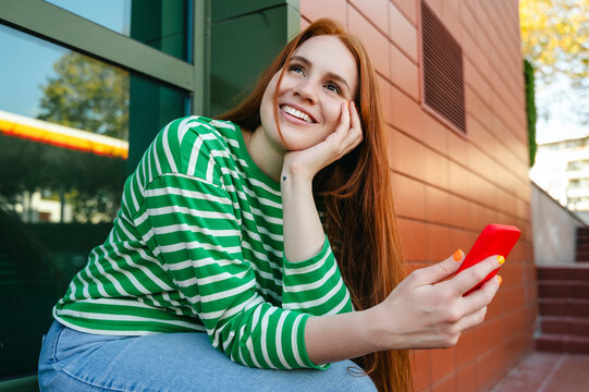 Happy redhead woman sitting with mobile phone near building