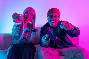 Young asian man and woman are playing video games together using controllers. They are sitting on the sofa in the living room with red and blue neon lighting. Couple gamer concept.