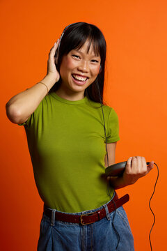 Happy woman listening to music through headphones and standing with stereo against orange background