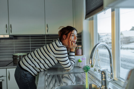 Woman leaning on kitchen counter looking out window at snowy landscape