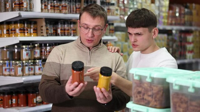 Emotional adult man and young guy, father and son, standing in supermarket aisle and discussing choice of aromatic natural spices in jars. High quality 4k footage