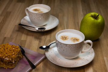 Two cups of cappuccino with spoons and an apple pie on plates ready to eat. A green apple lies next to it.