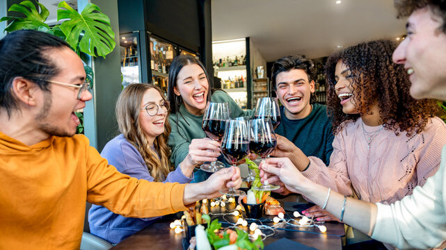 Group of multiracial friend toasting red wine sitting at bar restaurant table - Millennial people enjoying dinner party together - Life style concept with diverse guys and girls hanging out weekend