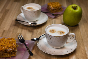 Two cups of cappuccino with spoons and an apple pie on plates ready to eat. A green apple lies next to it.
