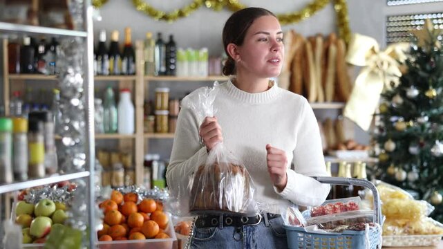 Enthusiastic young woman in casual sweater choosing traditional Italian fruitcake panettone while filling basket with treats for Christmas celebrations in grocery store decorated with holiday tinsel