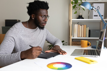 African American male graphic designer working at home using laptop and graphic tablet.