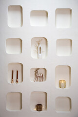 Minimalist wall display with built-in niches showcasing elegant holiday decor, including a white vase with delicate branches, metallic reindeer figurine, candles, and a glowing lantern.