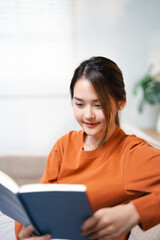 Naklejka premium girl enjoying an interesting novel reading in living room . Hobbies and activities. Happy young asian woman reading book, sitting on sofa lady enjoying interesting story, studying for exam,